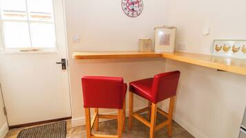 Una pequeña cocina con dos taburetes rojos, una encimera de madera, un reloj de pared con motivos de café y una ventana con persianas.