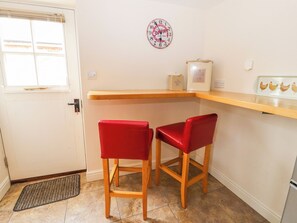 A small kitchen with two red bar stools, a wooden countertop, a coffee-themed wall clock, and a window with blinds. - The Shippon (Tarporley)