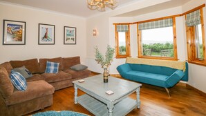 A living room with two sofas, a coffee table, and a chandelier. There are framed pictures on the wall and windows with wooden frames. - Ptarmigan (Grantown-on-Spey)