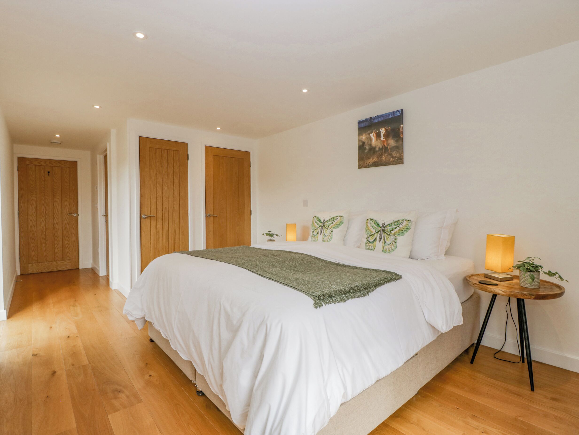 Une chambre à coucher avec un grand lit, un plancher en bois, deux portes, une table de chevet avec une lampe et un tableau accroché au mur.