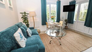 Ein Wohnzimmer mit einem petrolfarbenen Sofa, einem Glastisch, einem Esstisch mit Stühlen, einem an der Wand montierten Fernseher und einem großen Fenster mit Vorhängen.