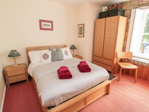 A neatly made bed with two red towels, a wooden wardrobe, a chair by the window, and a nightstand with a lamp. - Simonside Apartment (Morpeth)