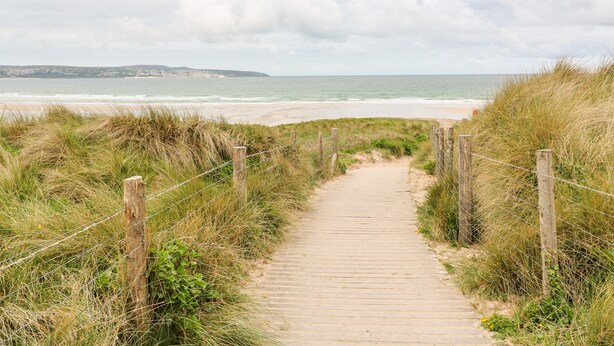 Un sendero de madera que lleva a una playa, con una cerca a un lado y césped al otro.