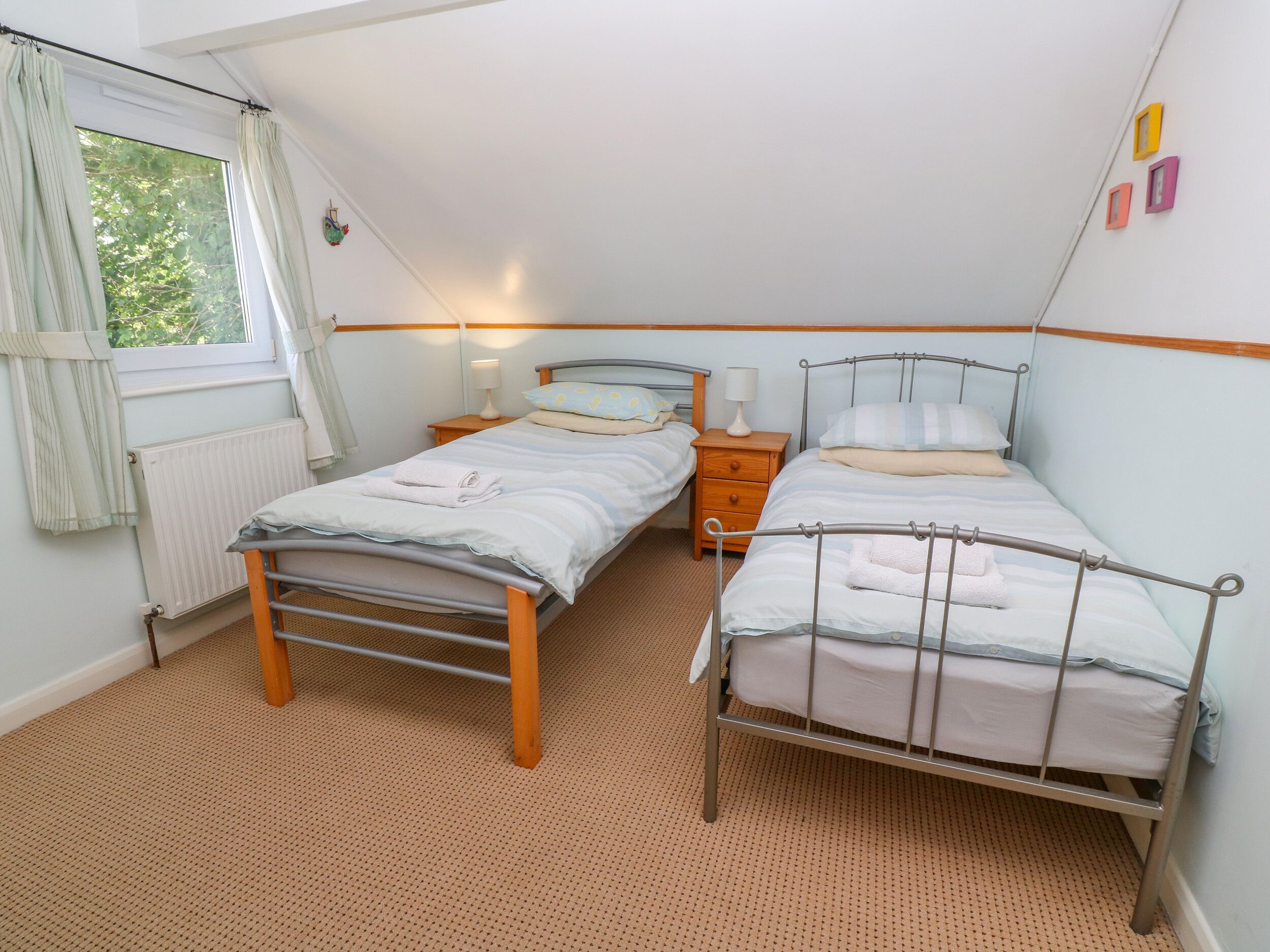 Une chambre avec deux lits simples, une fenêtre avec des rideaux, un radiateur et une table de chevet en bois avec une lampe.
