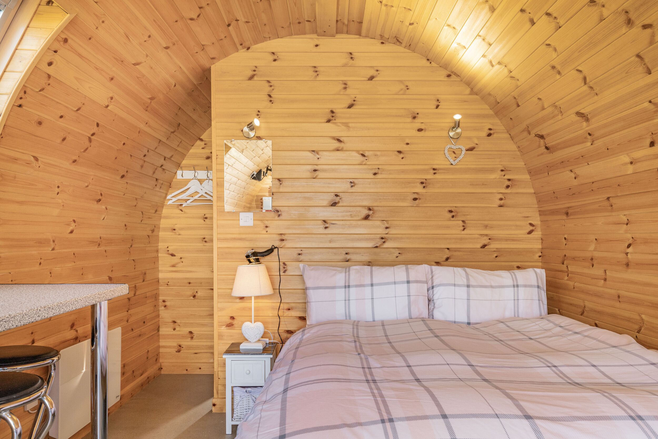 A cozy bedroom with a wooden ceiling, a bed with a plaid duvet, a bedside table with a lamp, and a small nightstand.
