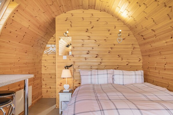 A cozy bedroom with a wooden ceiling, a bed with a plaid duvet, a bedside table with a lamp, and a small nightstand. - Two Hoots (Flint)