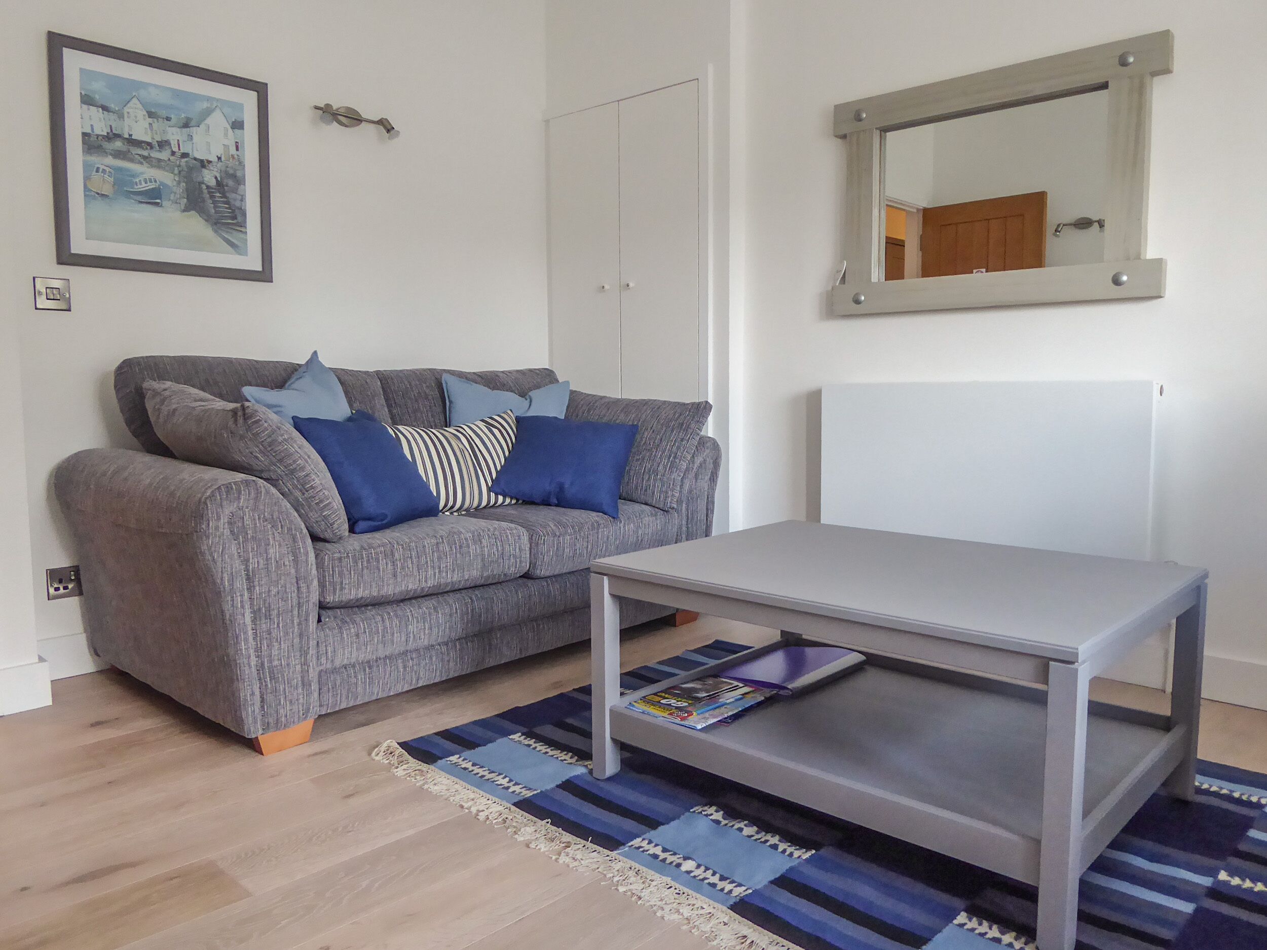 Un salon avec un canapé gris, des coussins bleus, une table basse blanche, un tapis rayé bleu et beige, un tableau encadré et un miroir au mur.