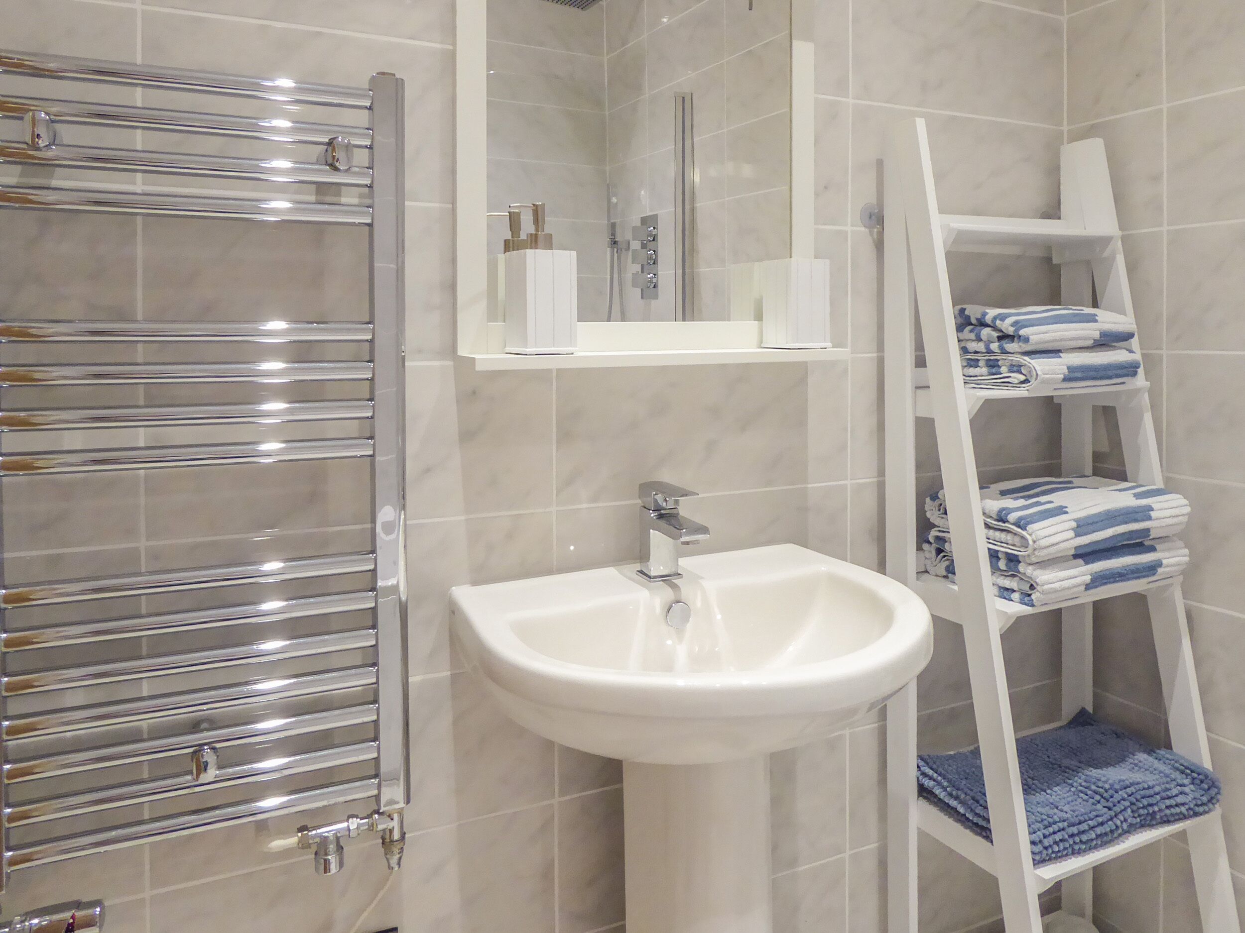 A bathroom with a white sink, a chrome towel rack, a white shelf with toiletries, and a white towel rack with towels.