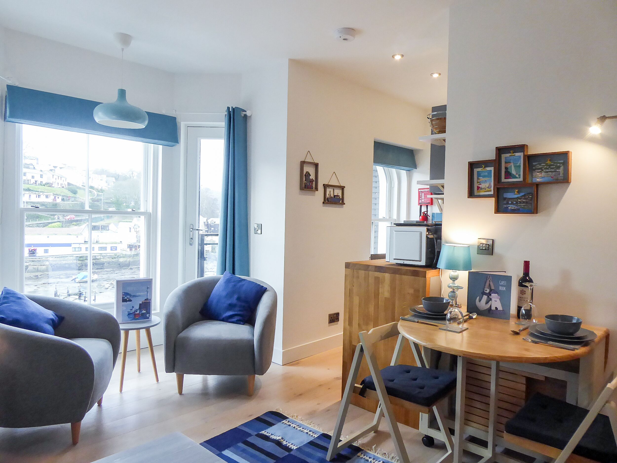 Un salon moderne comprenant un canapé, un fauteuil et une table à manger, avec une vue dégagée sur l’extérieur grâce à une grande fenêtre, ainsi qu’un coin cuisine équipé d’un four à micro-ondes et agrémenté de tableaux encadrés.
