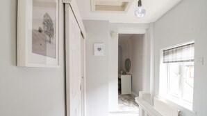 A hallway with a white door, a framed picture, a pendant light, and a window with blinds. - End Cottage (Ashbourne)