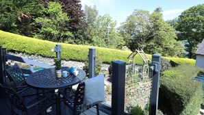 A deck with a round table and chairs, surrounded by a stone wall and lush greenery. - Bryn Teg Cottage (Menai Bridge)