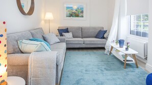 A living room with a grey sofa, a blue rug, a small table with a lamp, and a window with curtains. - Bryn Teg Cottage (Menai Bridge)