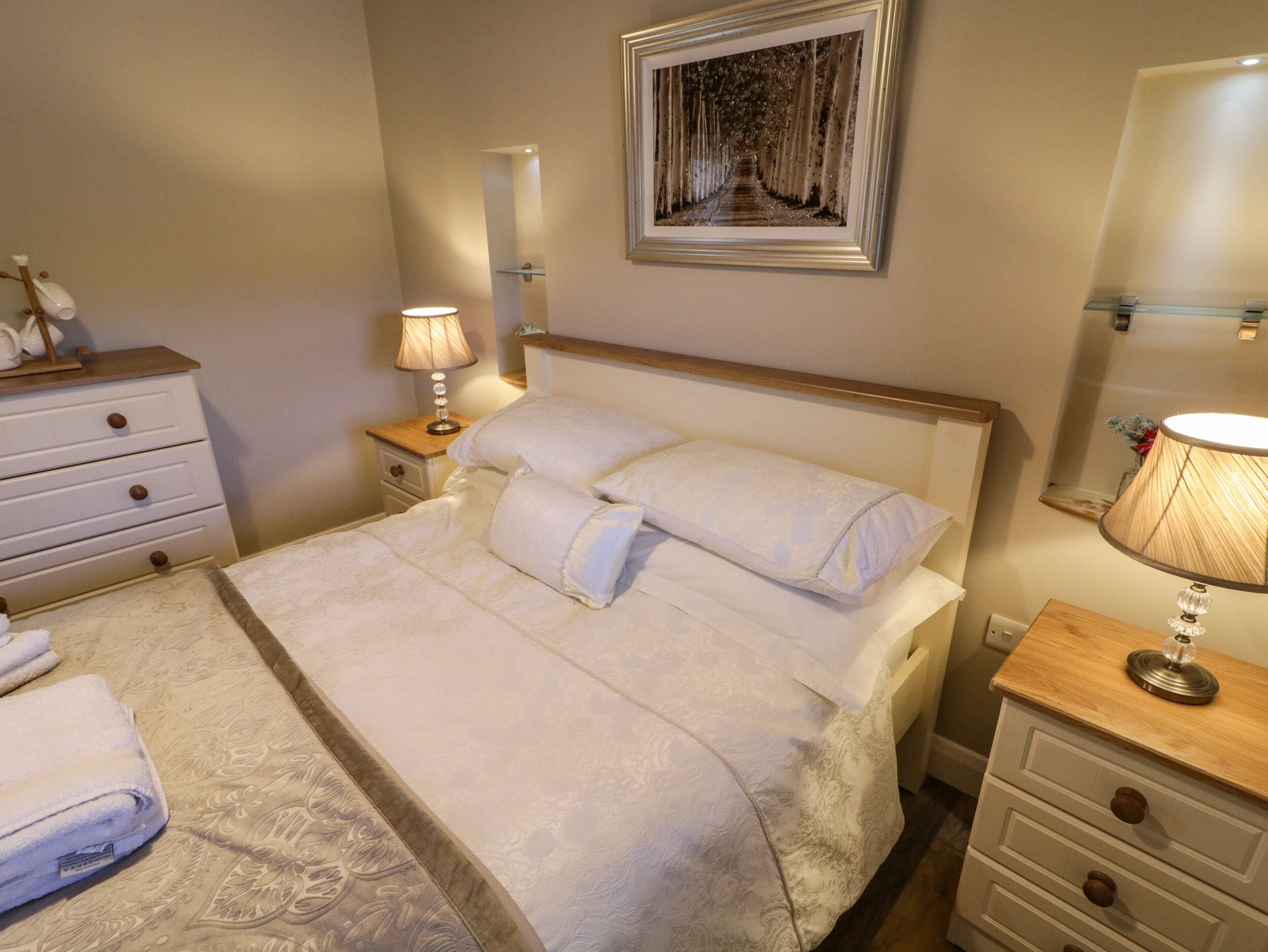 A neatly made bed with a white comforter and pillows, a bedside table with a lamp, a nightstand with a lamp, and a wall-mounted picture.