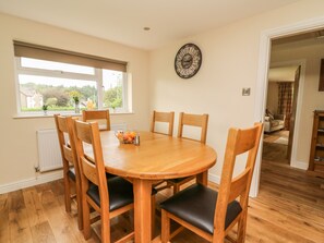 A dining area with a round wooden table, six wooden chairs with black cushions, a metal fruit bowl, and a wall clock.