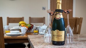 A bottle of Taittinger champagne with two glasses attached, placed on a marble table in a dining area with a fruit bowl and cups in the background. - New Peny (Llandrindod Wells)