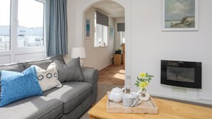 Un salon moderne avec un canapé gris, une table basse en bois sur laquelle se trouve un plateau avec du thé et des tasses, une cheminée et une vue sur l’extérieur grâce à de grandes fenêtres.