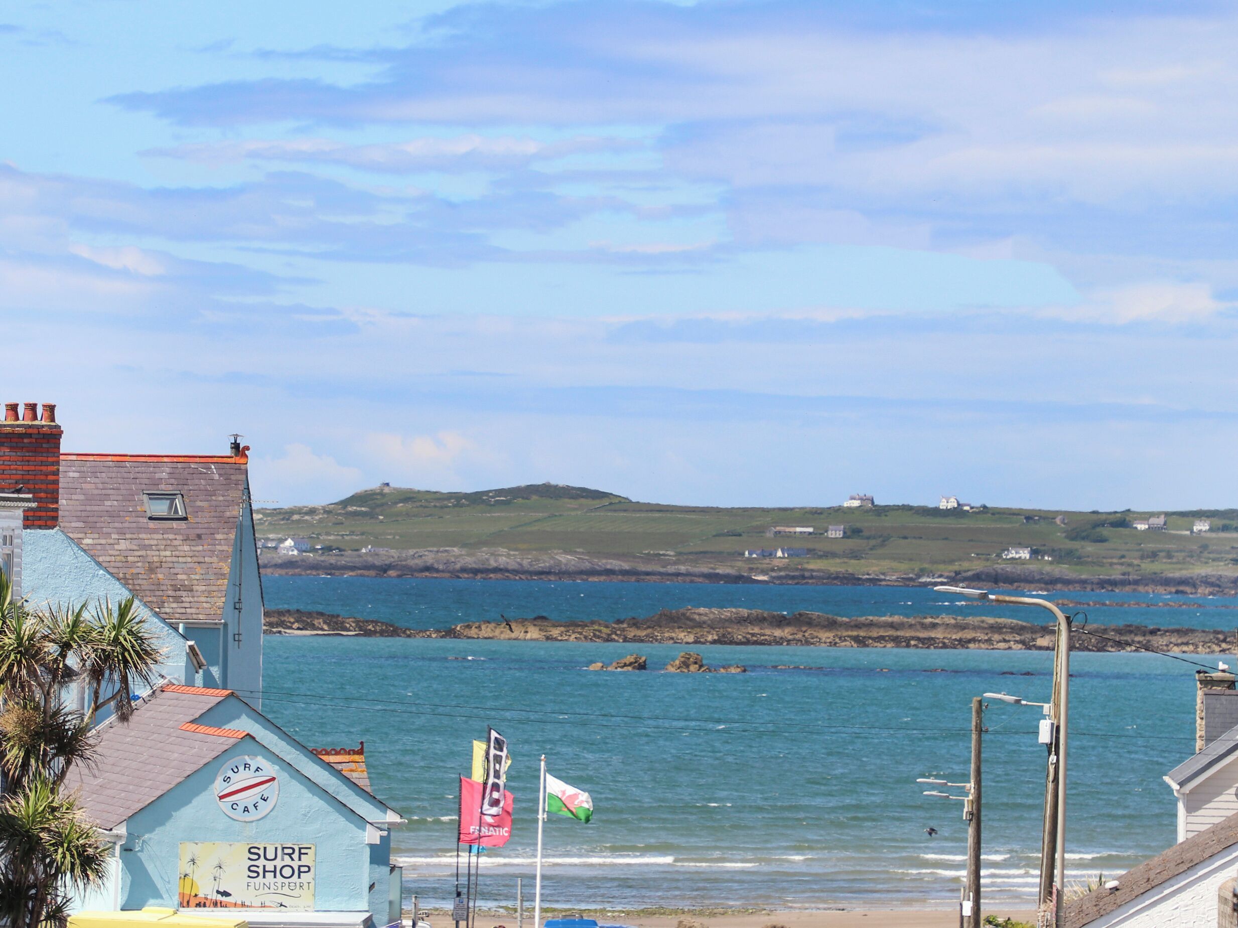 Un edificio costero con un letrero que dice 'Surf Shop', vista al mar y una colina con casas al fondo.