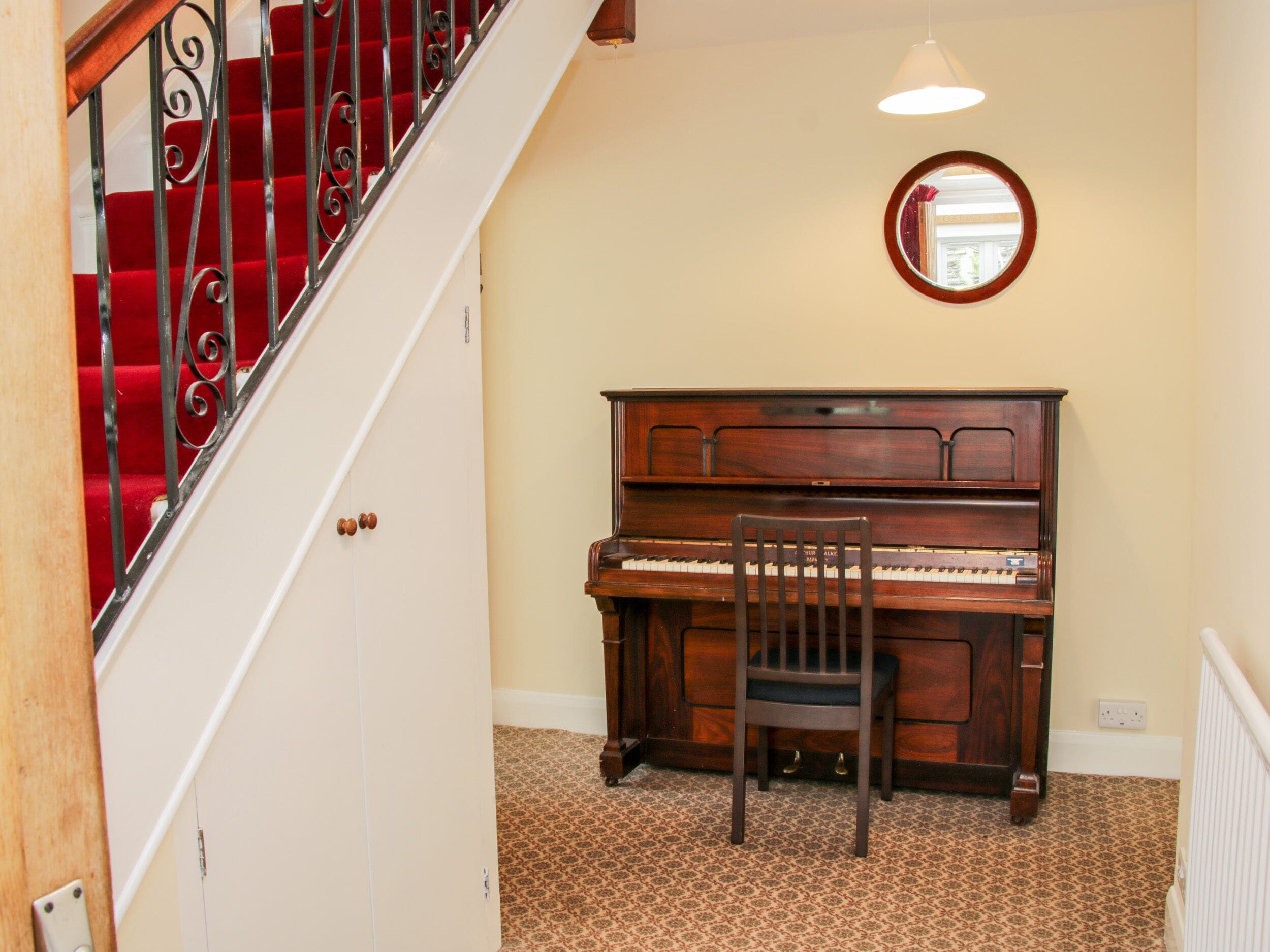 Una habitación con un piano de cola, una silla de madera, una escalera con alfombra roja y un espejo redondo en la pared.