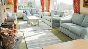 Una sala de estar acogedora con dos sofás, una mesa de centro y una alfombra. Hay ventanas con cortinas, una lámpara y un cesto de leña.