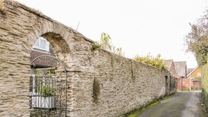 Un muro de piedra con una reja metálica, un camino estrecho y edificios residenciales al fondo.
