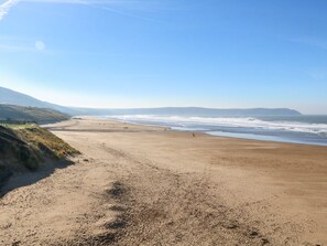A wide sandy beach with gentle waves, a clear sky, and distant hills.