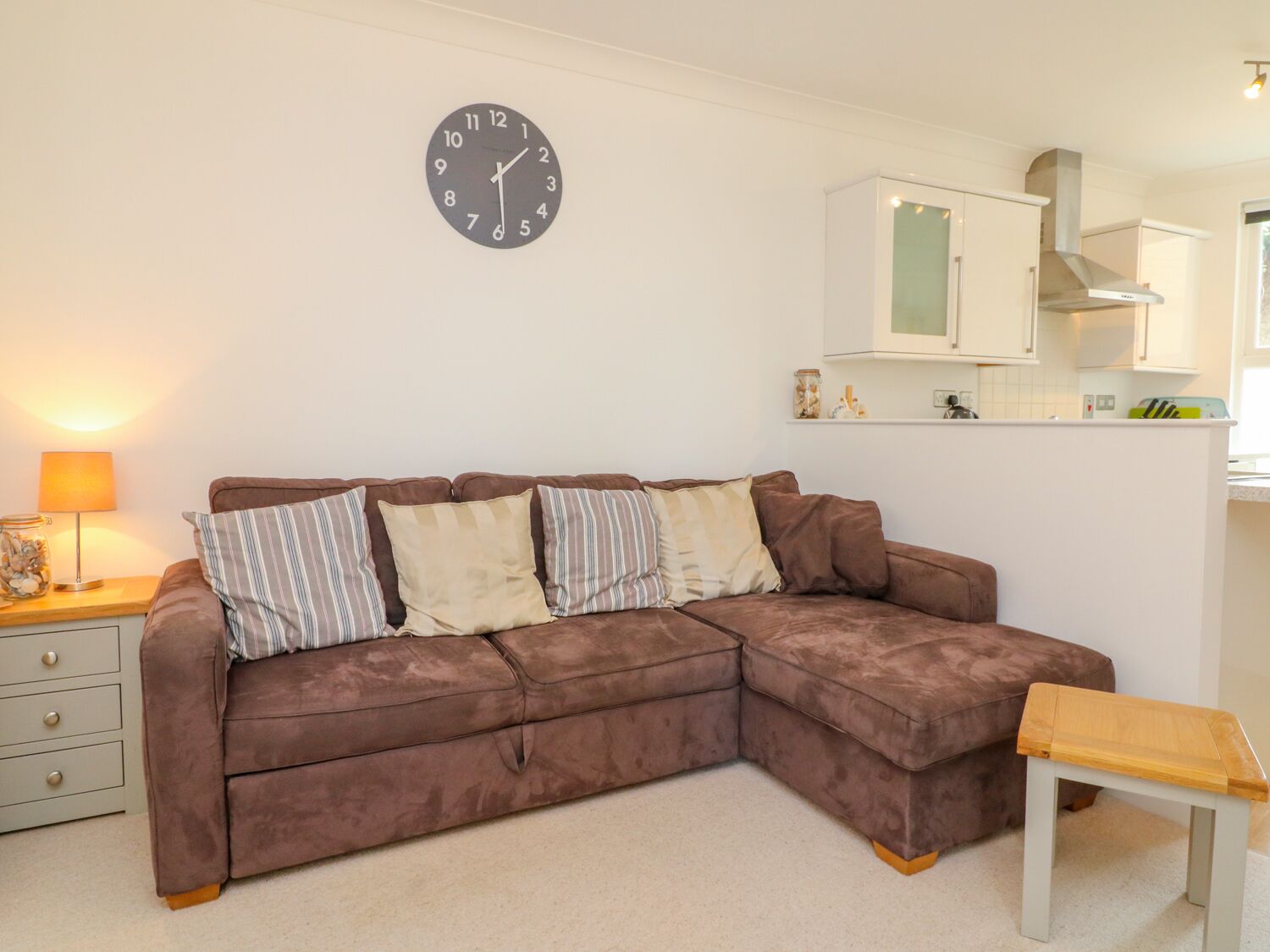 A living room with a brown sectional sofa, a wooden coffee table, a wall clock, and a kitchen area with white cabinets and a stove.