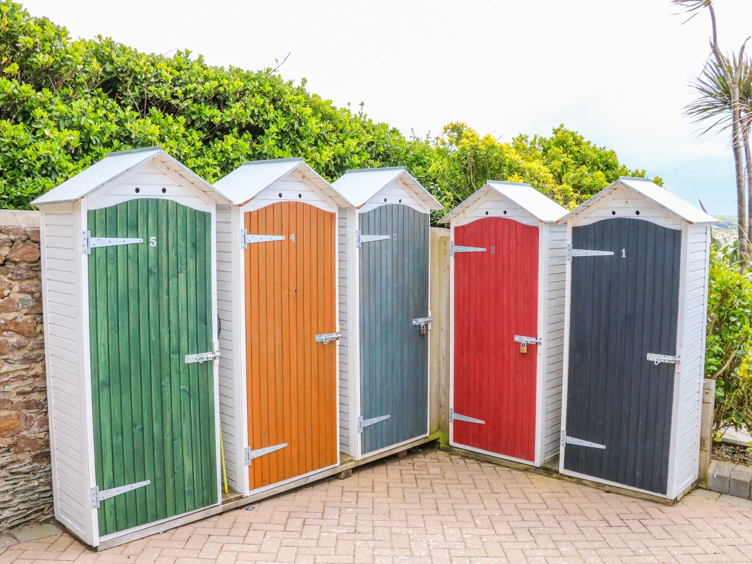 Cinco cabinas de ducha exteriores portátiles con puertas numeradas, cada una de un color diferente, alineadas contra una pared de piedra con un seto al fondo.