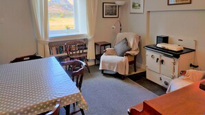 A cozy room with a dining table, a chair, a bookshelf, a window with curtains, a lamp, a stove, and a bed. - Ornum (Thurso)