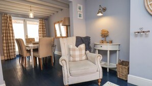 A living room with a beige armchair, a dining table with chairs, a lamp, and a small side table with decorative items. - Adventure Cottage (Whitby)