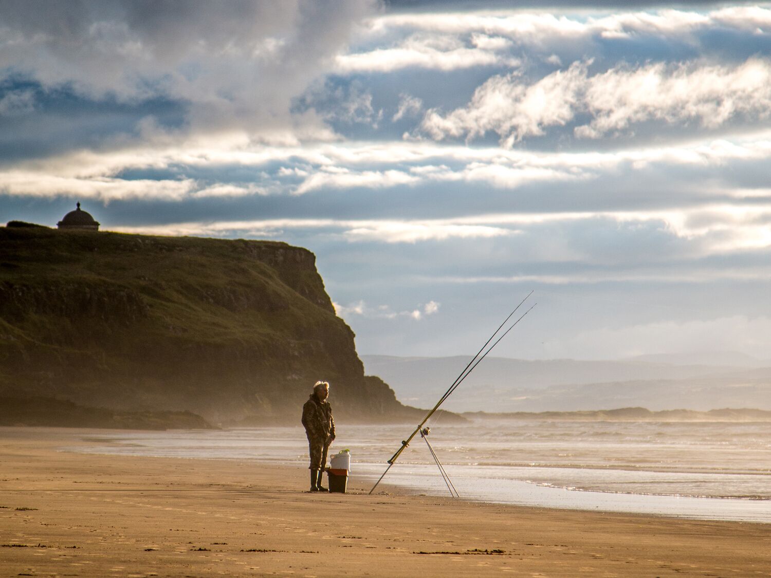 A person fishing on a sandy beach with a cliff and a dome structure in the background.