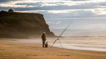 A person fishing on a sandy beach with a cliff and a dome structure in the background.