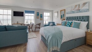A spacious bedroom with a large bed, a desk with a chair, a sofa, and a view of the sea through the windows. - 6 The Beaches (Weymouth)
