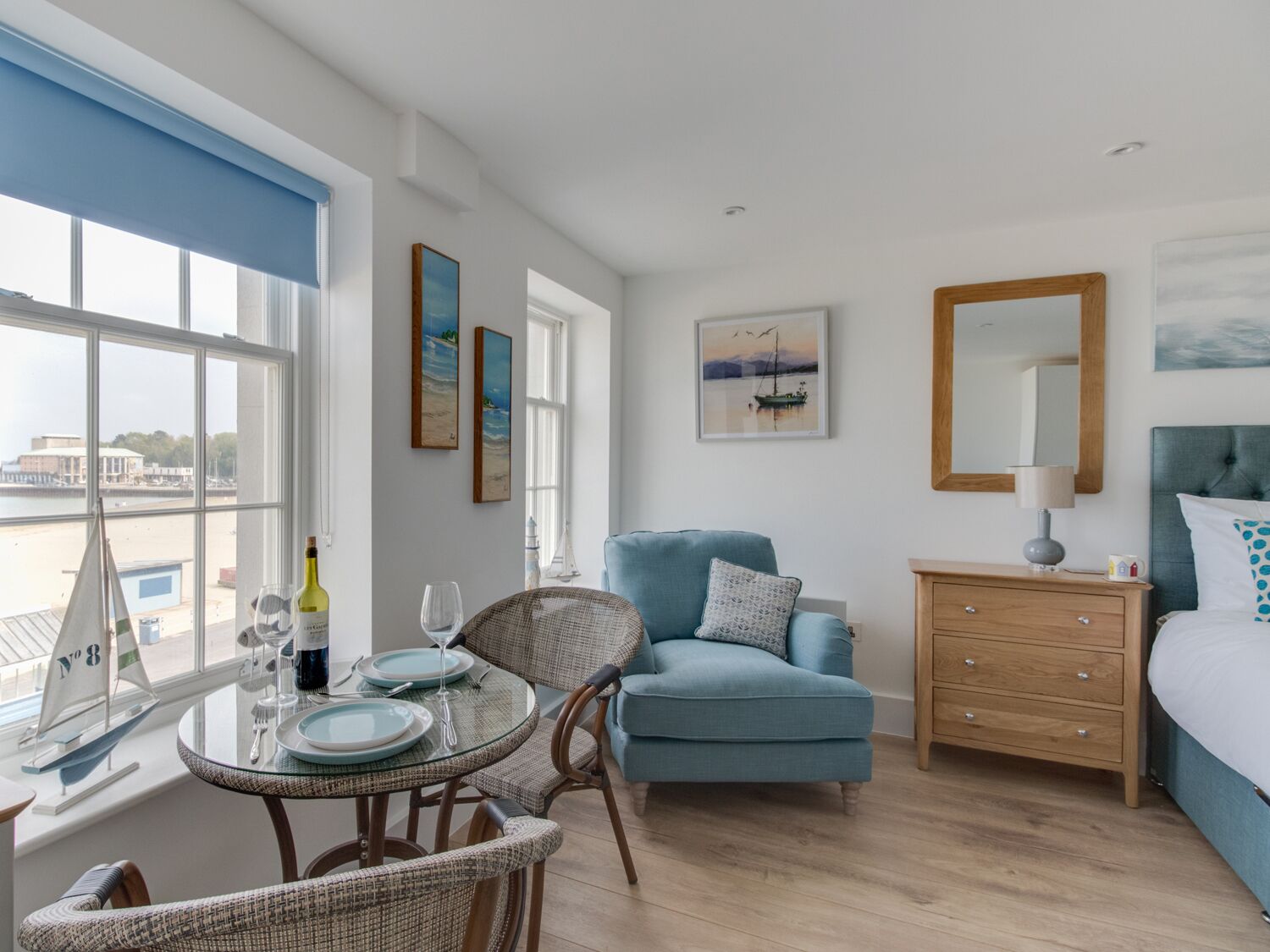 A room with a bed, a dresser, a chair, a table with plates and glasses, and a sailboat model by the window.