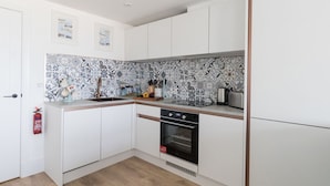 A compact kitchen with white cabinets, a patterned backsplash, a built-in oven, and a small countertop with various items. - 6 The Beaches (Weymouth)