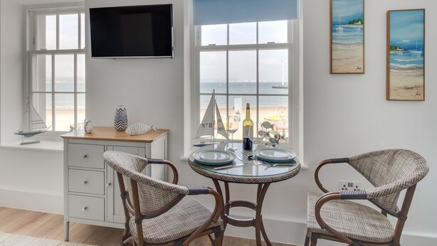 A dining area with wicker chairs, a round glass table, and a view of the sea through large windows. - 6 The Beaches (Weymouth)