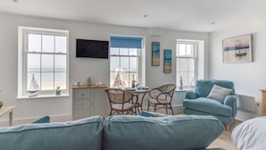 A modern living room with a sofa, a dining table, and chairs. There are windows with a view of the sea. - 6 The Beaches (Weymouth)