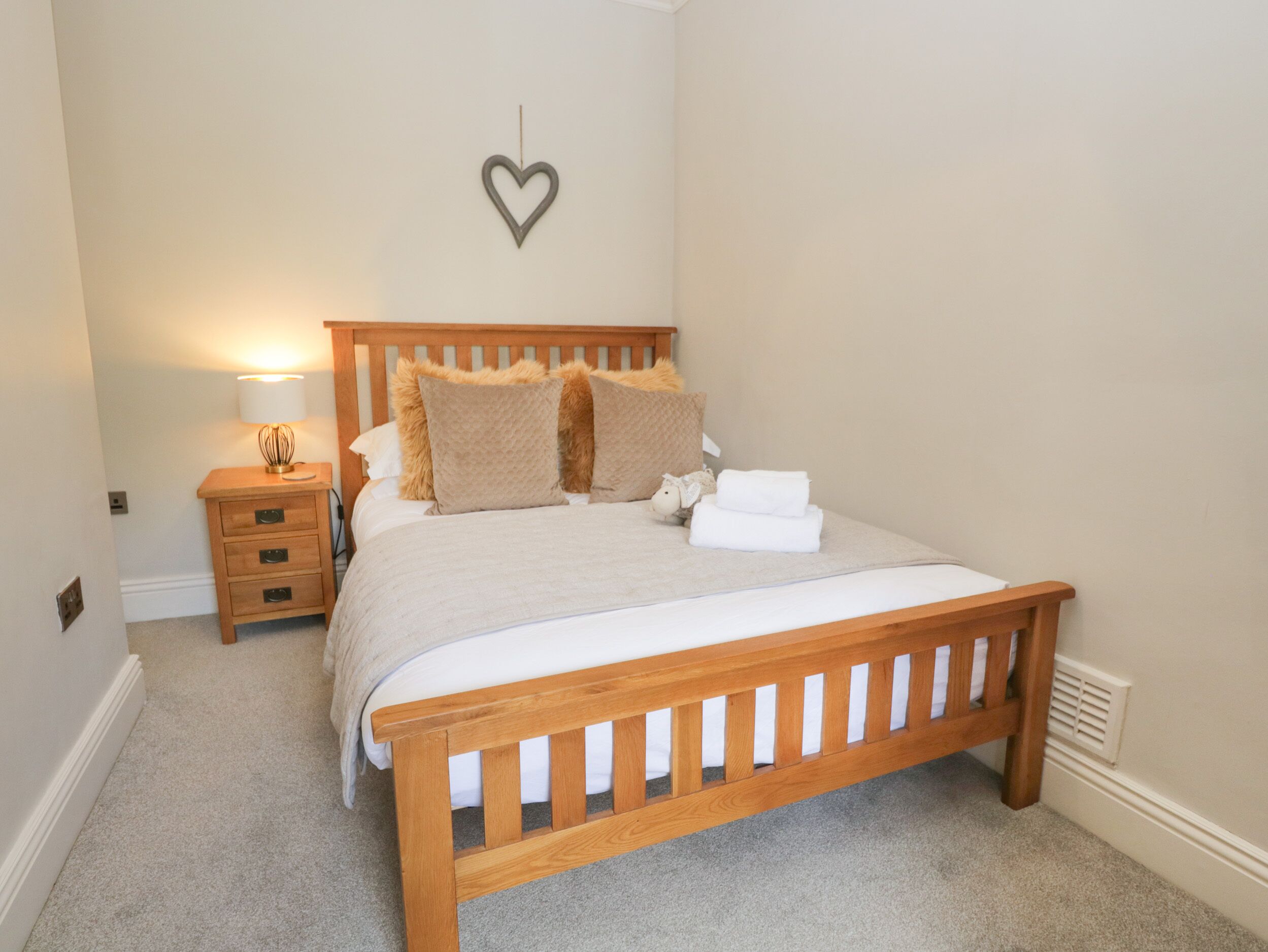 A bedroom with a wooden bed, a nightstand, a lamp, and a heart-shaped decoration on the wall.