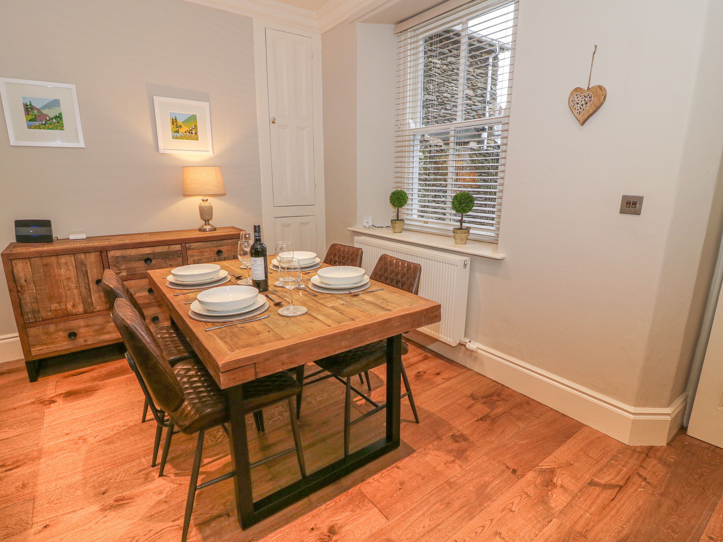 A dining area with a wooden table set for four, a bench seating, a radiator, a window with blinds, and a wall-mounted light fixture.