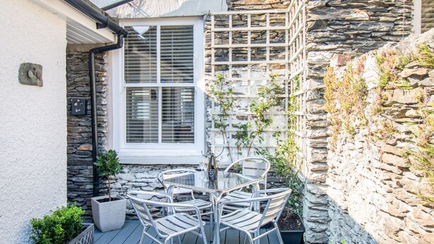 A patio with a table and chairs, a window with blinds, and a stone wall with climbing plants. - Little Herdwick (Windermere)