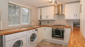 A compact kitchen with a washing machine, oven, and a window with blinds. - Little Herdwick (Windermere)