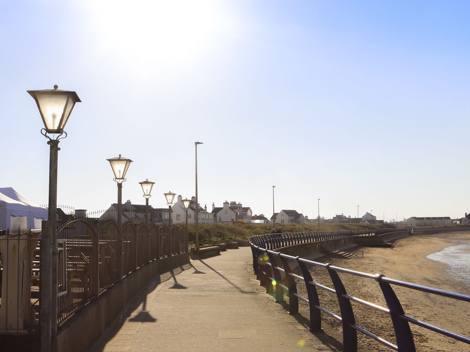 Un paseo marítimo con farolas, una barandilla de metal y una playa arenosa.