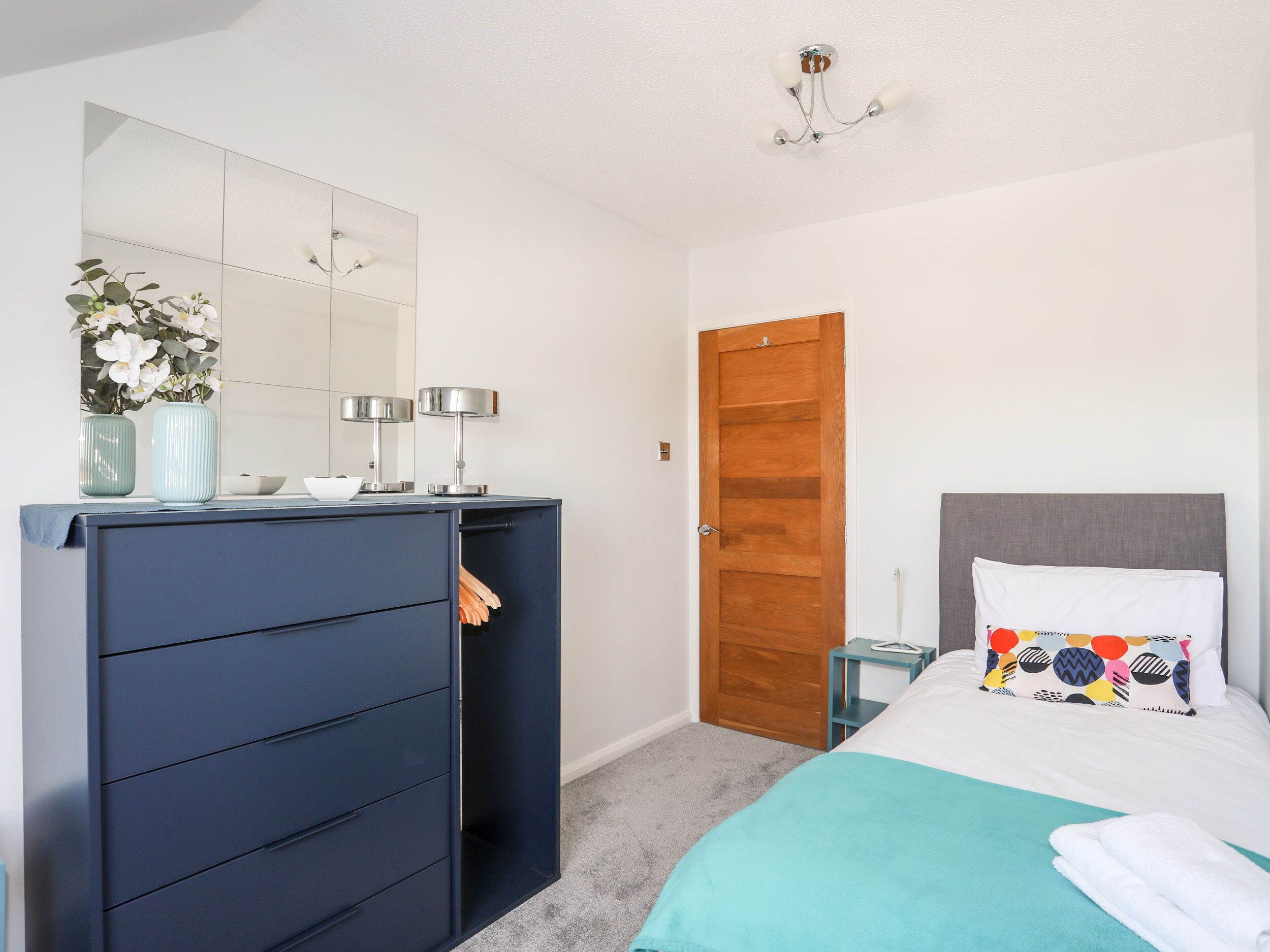 Een slaapkamer met een bed, een dressoir, een spiegel, een deur en een kleine tafel met een lamp.