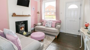 A cozy living room with a fireplace, a sofa, a pink ottoman, and a television on the wall.