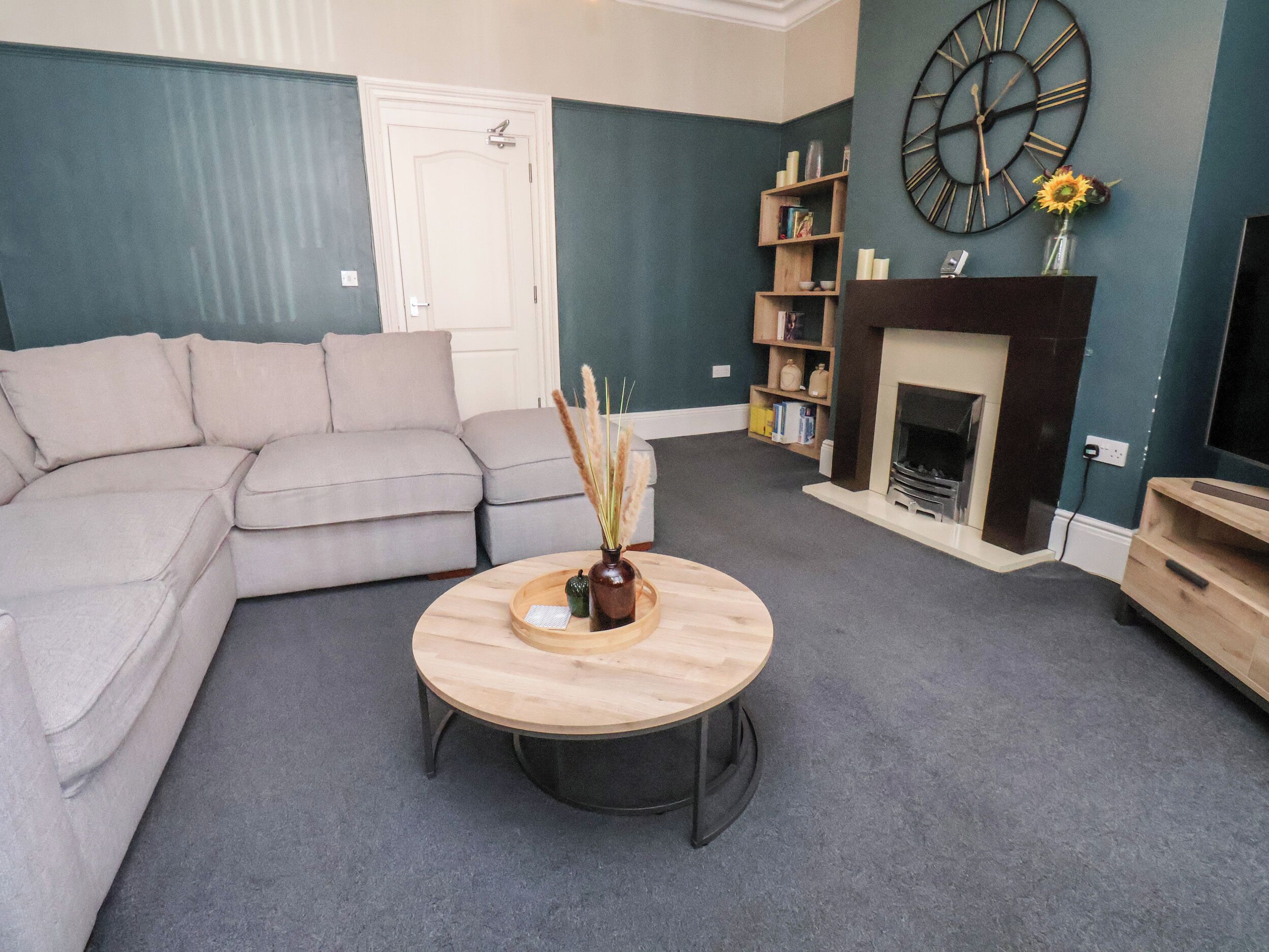 A living room with a sectional sofa, a round coffee table, a bookshelf, a fireplace, and a wall clock.