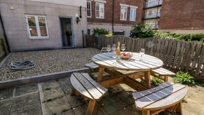 A wooden outdoor patio with a round table and benches, surrounded by a gravel area and a wooden fence.