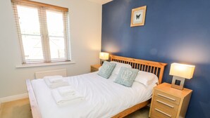 A neatly made bed with white linens and folded towels, a wooden headboard, two bedside tables with lamps, and a window with blinds.