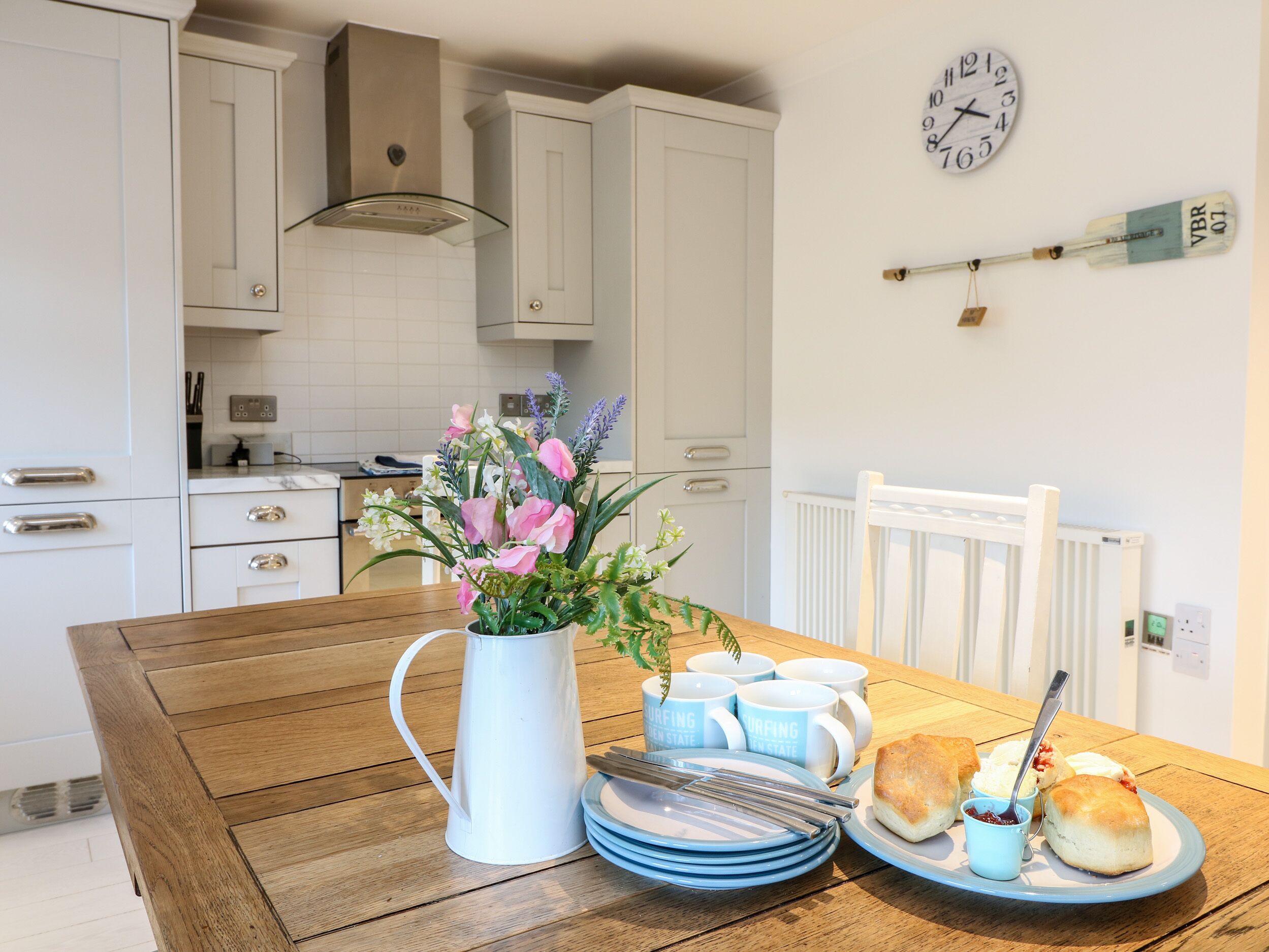 A kitchen with white cabinets, a wooden table, a white pitcher, blue mugs, and a plate with scones and jam.