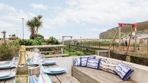 Ein Holztisch, gedeckt mit Tellern, Gläsern und einer Weinflasche, mit Blick auf eine malerische Küstenlandschaft, inklusive einer Holzbank und Kissen.