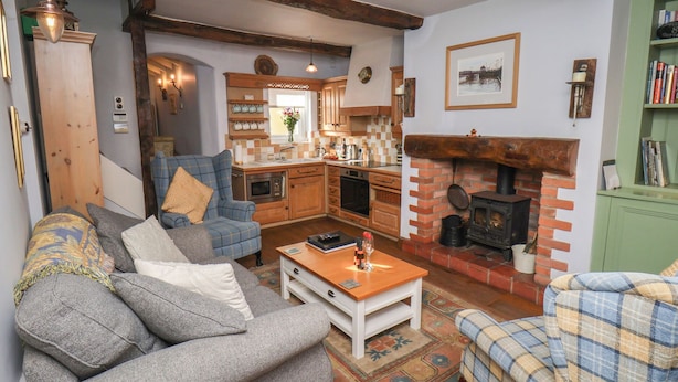 A cozy living room with a fireplace, a sofa, a coffee table, and a kitchen area with wooden cabinets and a brick stove. - Cobbles House (Whitby)
