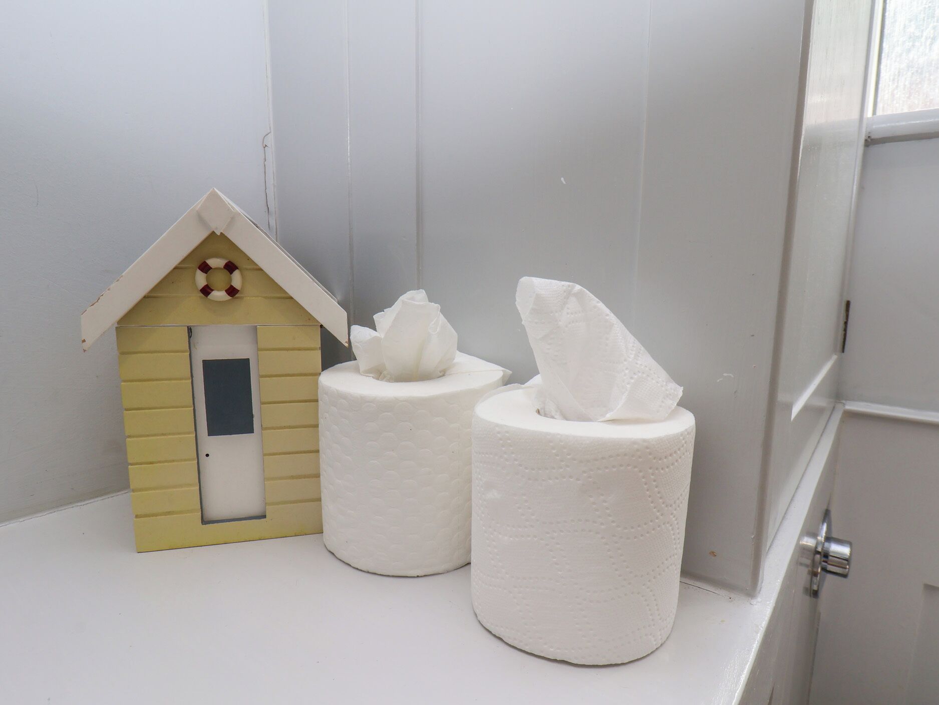 Een klein, geel huis met een reddingsboei, twee toiletrollen en een witte deur.